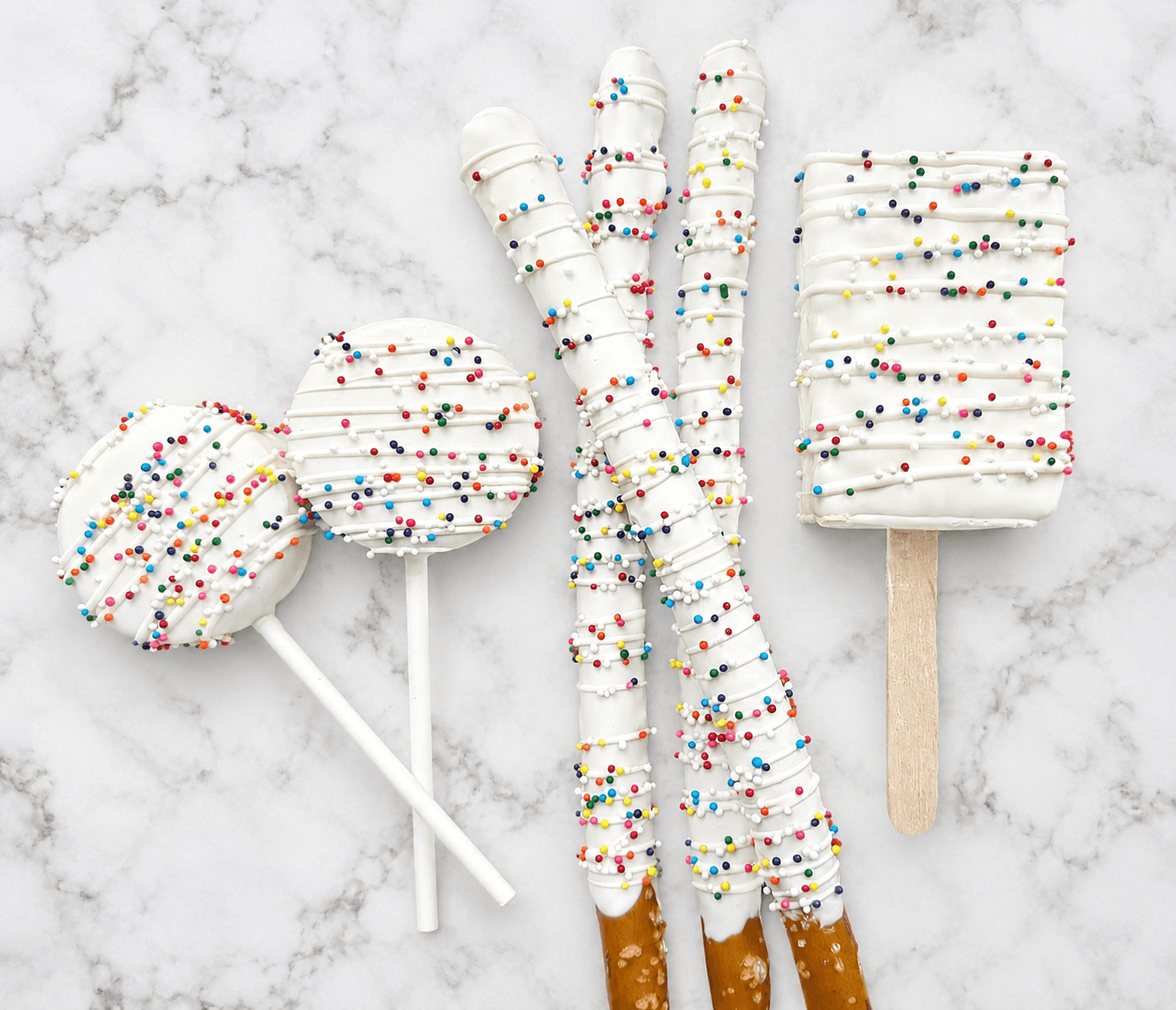 White chocolate-dipped cake pops, pretzel rods, and a cakesicle covered in rainbow nonpareil sprinkles